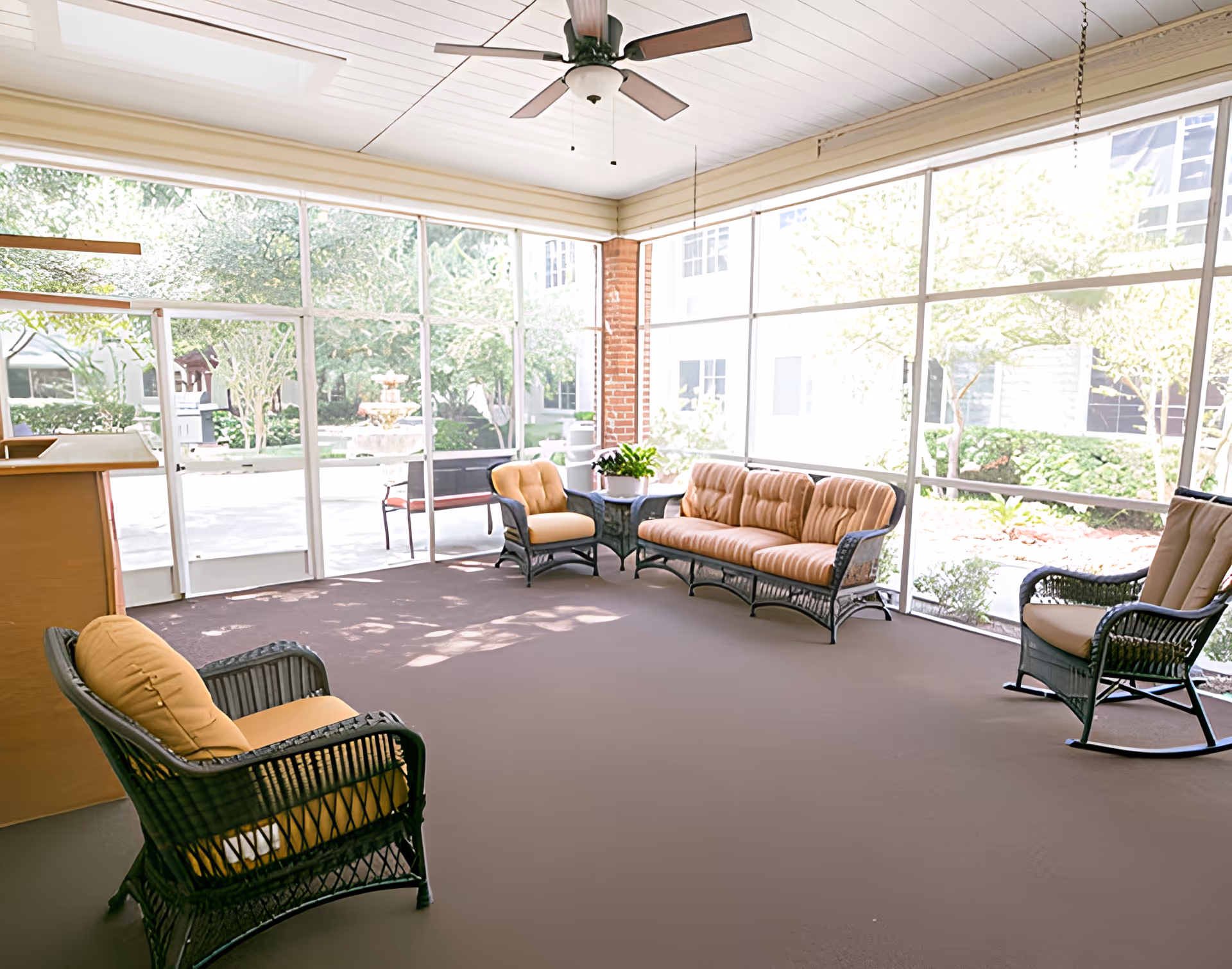 A bright sunroom with large windows overlooking a garden area. The room contains wicker furniture with yellow cushions, including a sofa, two armchairs, and a rocking chair. A ceiling fan is mounted on the white paneled ceiling.