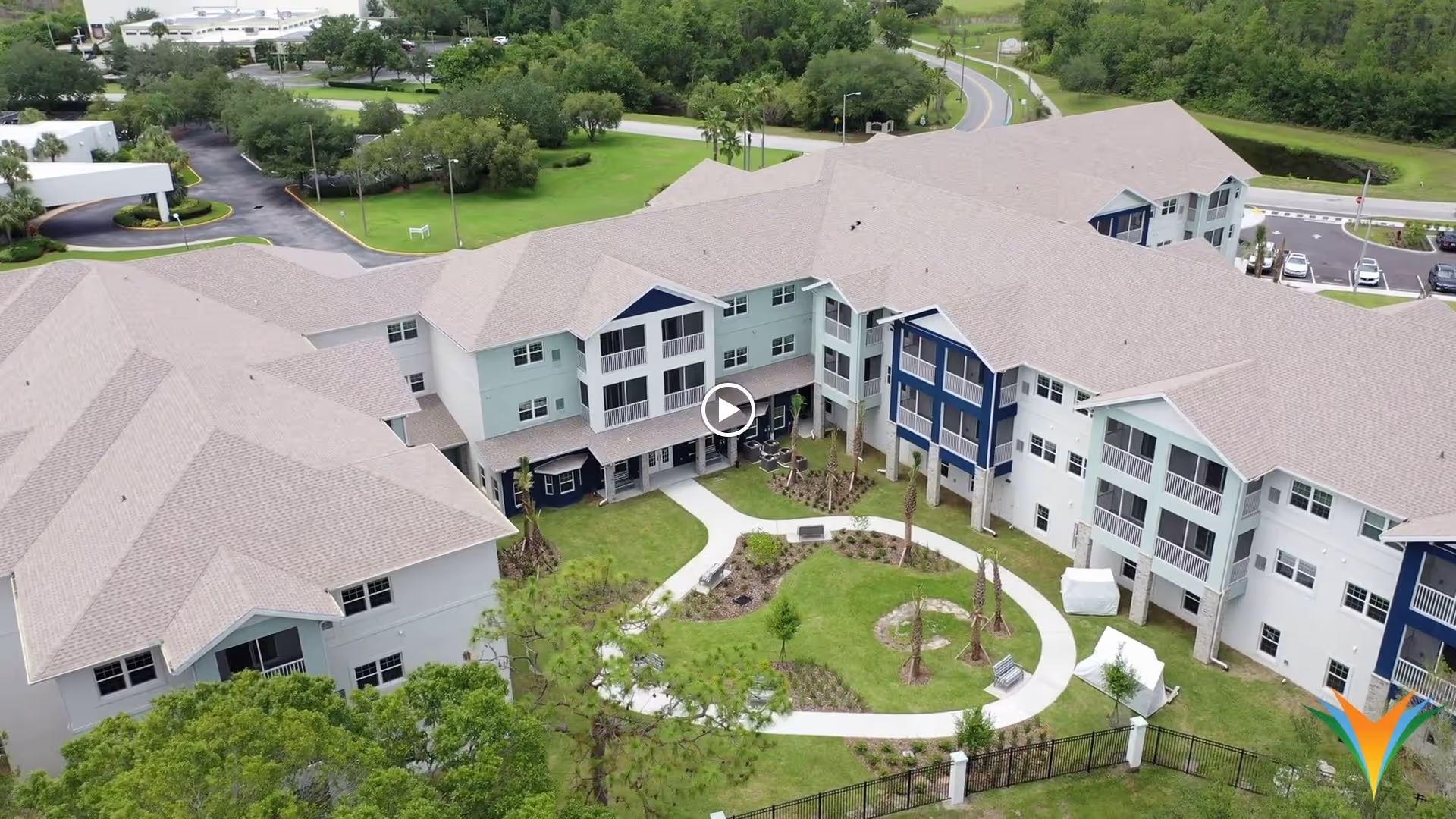 Aerial view of a senior living facility named Sunflower Springs at Trinity, showing a large multi-story building with beige roofs and white and blue exterior walls. The building surrounds a landscaped courtyard with walking paths, small trees, and benches. There are parking areas and roads visible in the background, along with green lawns and trees.