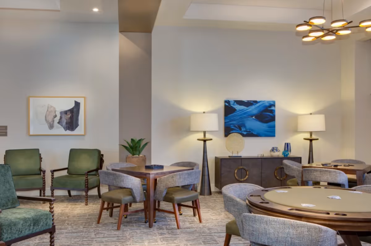 Communal seating area with round game tables, upholstered chairs, decorative lamps, artwork, and a sideboard.
