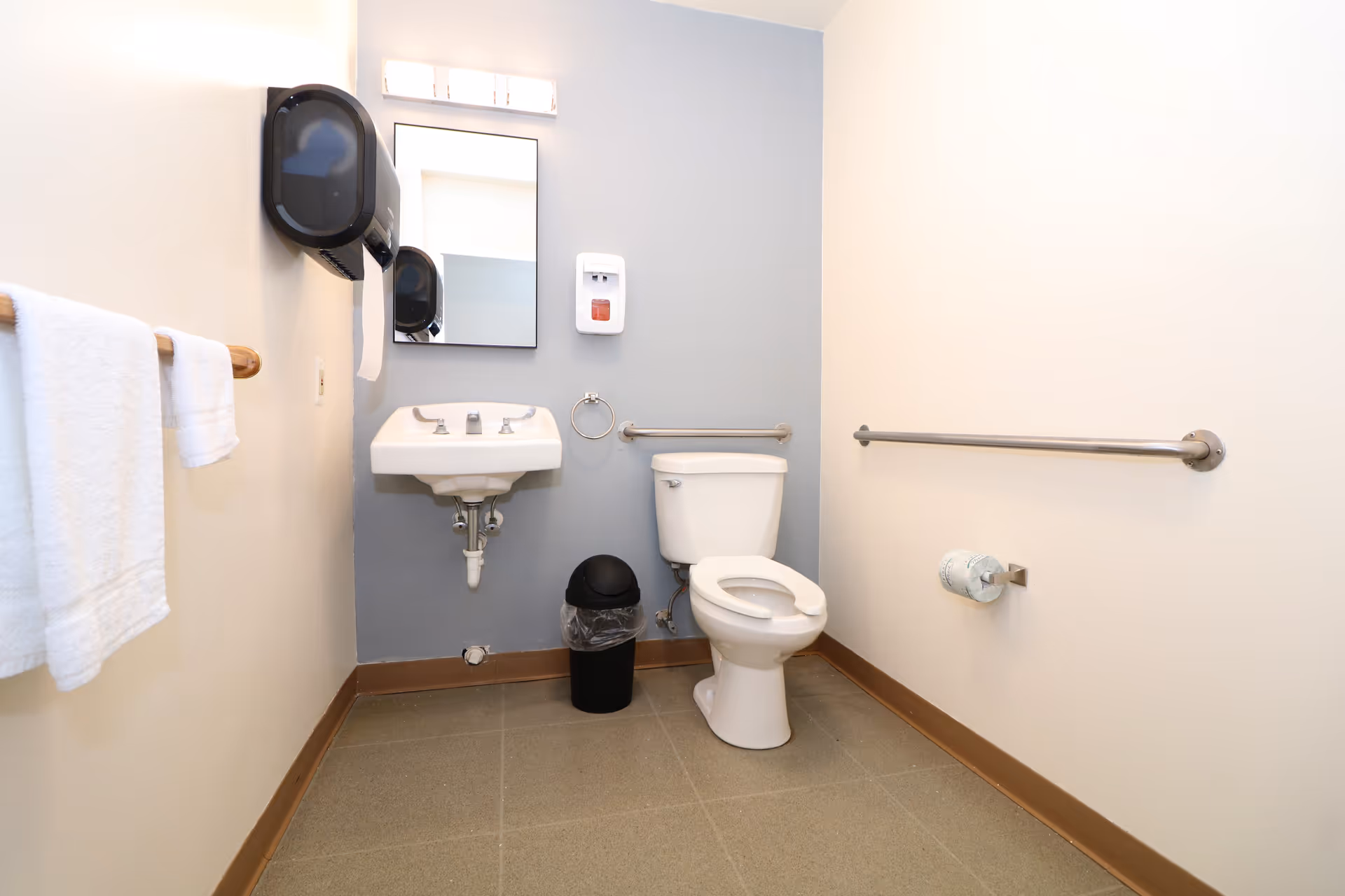 A clean, accessible bathroom with a white toilet, a wall-mounted sink, a mirror above the sink, a black trash can, a towel rack with white towels, a soap dispenser, and grab bars on the walls for support.