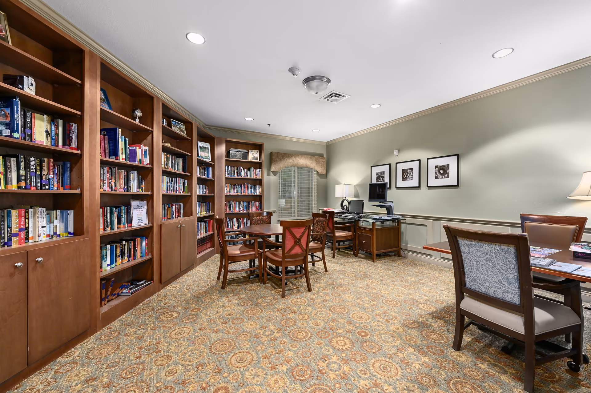 Interior view of a library or reading room with wooden bookshelves filled with books along the left wall, several wooden tables and chairs arranged in the center and right side of the room, a computer desk with a monitor and printer against the far wall, and framed pictures hanging on the wall above the desk. The room has patterned carpet flooring and recessed ceiling lights.