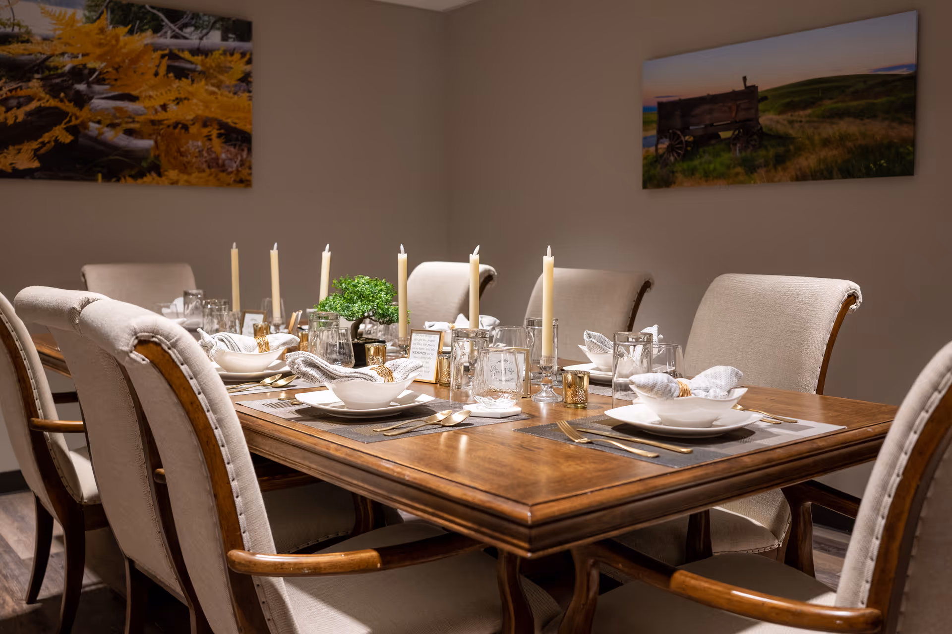 A dining room with a wooden table set for six people. The table is decorated with white plates, bowls, cloth napkins, glassware, gold-colored cutlery, tall white candles, and a small green plant centerpiece. Beige upholstered chairs surround the table. Two landscape paintings hang on the beige walls.