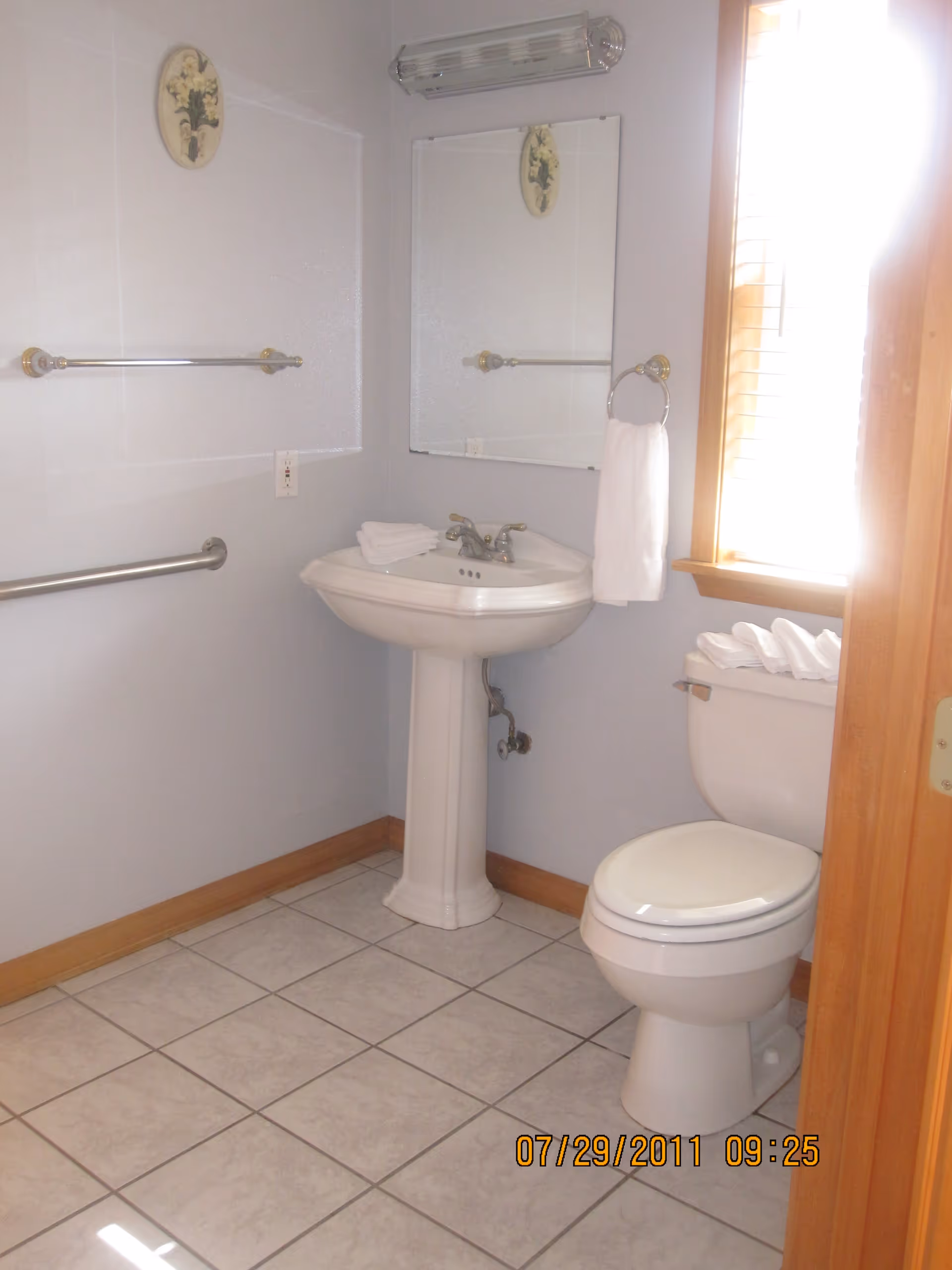A clean bathroom with a white pedestal sink, a toilet with folded towels on top, a large mirror above the sink, a towel ring with a white hand towel, and two towel bars on the wall. The floor is tiled, and there is a window with wooden blinds letting in bright light.
