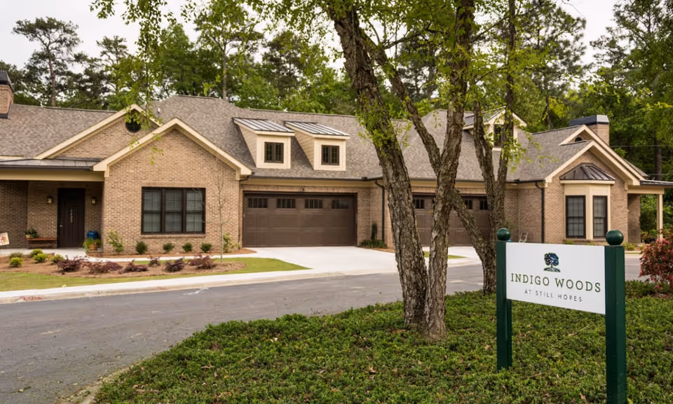 Exterior view of a brick building with multiple garage doors and windows, surrounded by trees and greenery. A sign in the foreground reads 'Indigo Woods at Still Hopes.'