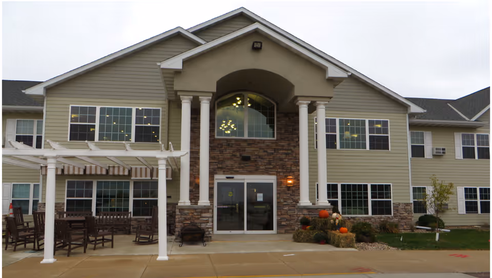 Front exterior view of a two-story senior living facility building with beige siding and stone accents. The entrance features double glass doors framed by four white columns supporting a small portico. To the left, there is a white pergola with several wooden chairs and tables underneath. On the right side near the entrance, there are hay bales decorated with pumpkins and flowers. The sky is overcast.