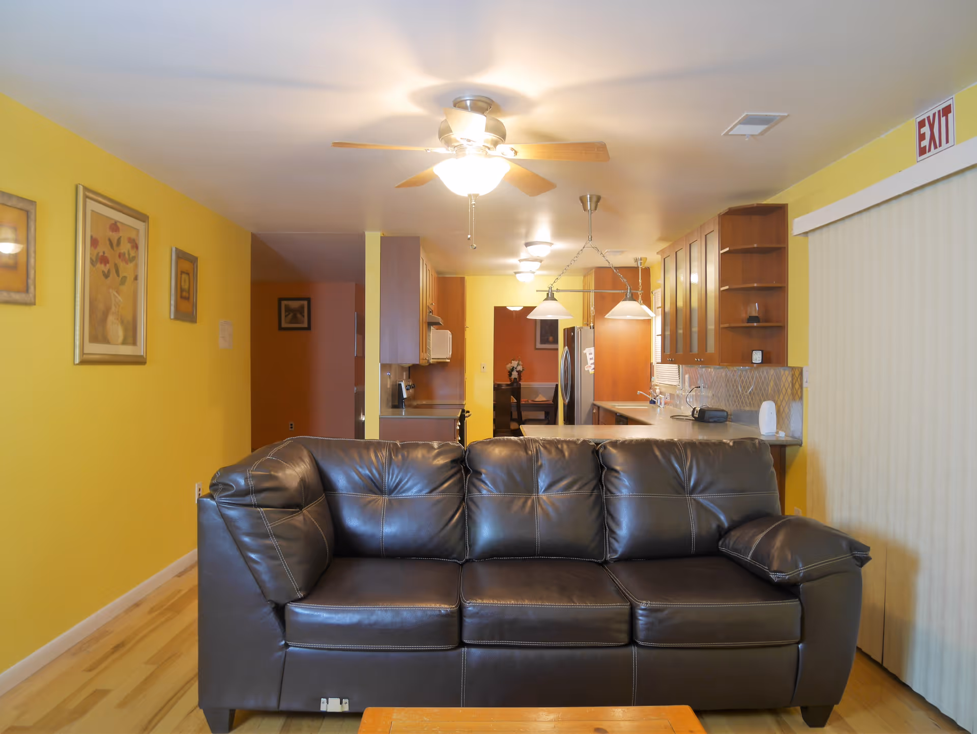 A dark leather sofa in a living room facing an open kitchen with yellow walls, hanging lights and a ceiling fan.
