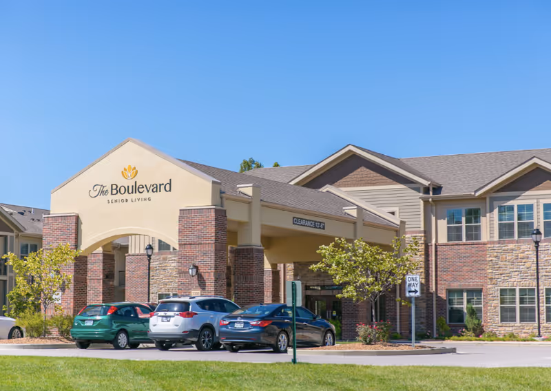 Exterior view of The Boulevard Senior Living facility showing the entrance with a covered drop-off area, brick and stone facade, several parked cars, and clear blue sky.