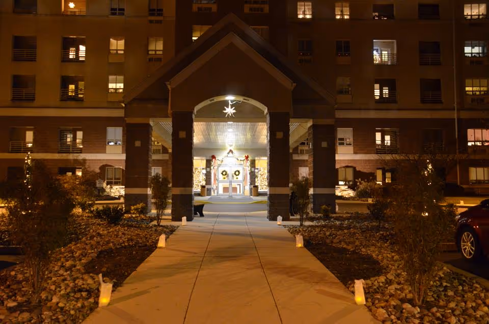 Night view of a lit covered entryway and walkway leading to the decorated front doors of a multi-story senior living building.