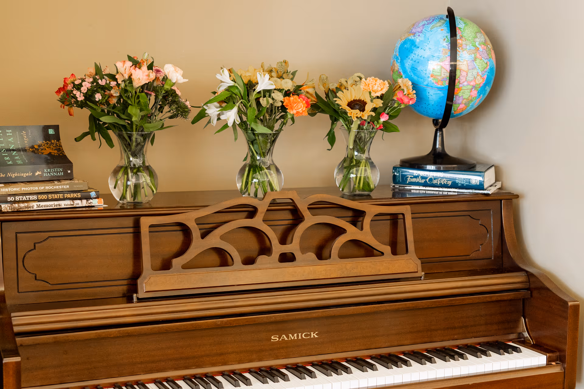 A wooden upright piano with three glass vases of colorful flowers on top, along with a globe and several stacked books, set against a beige wall.