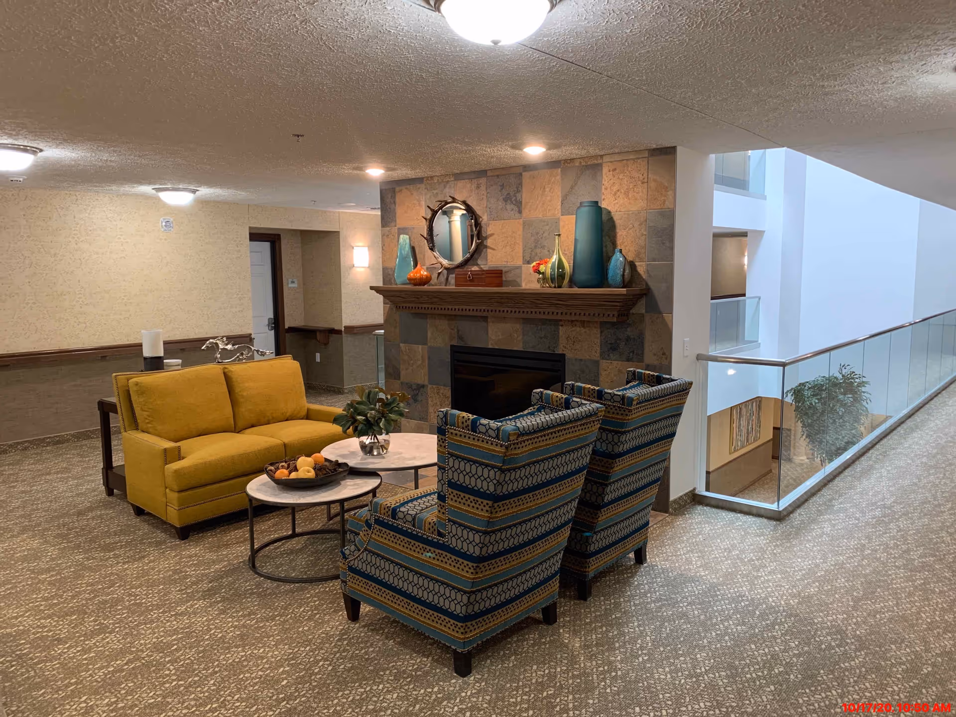 A lounge area with a yellow loveseat, patterned armchairs, coffee tables and a decorative tiled fireplace in a retirement facility corridor.