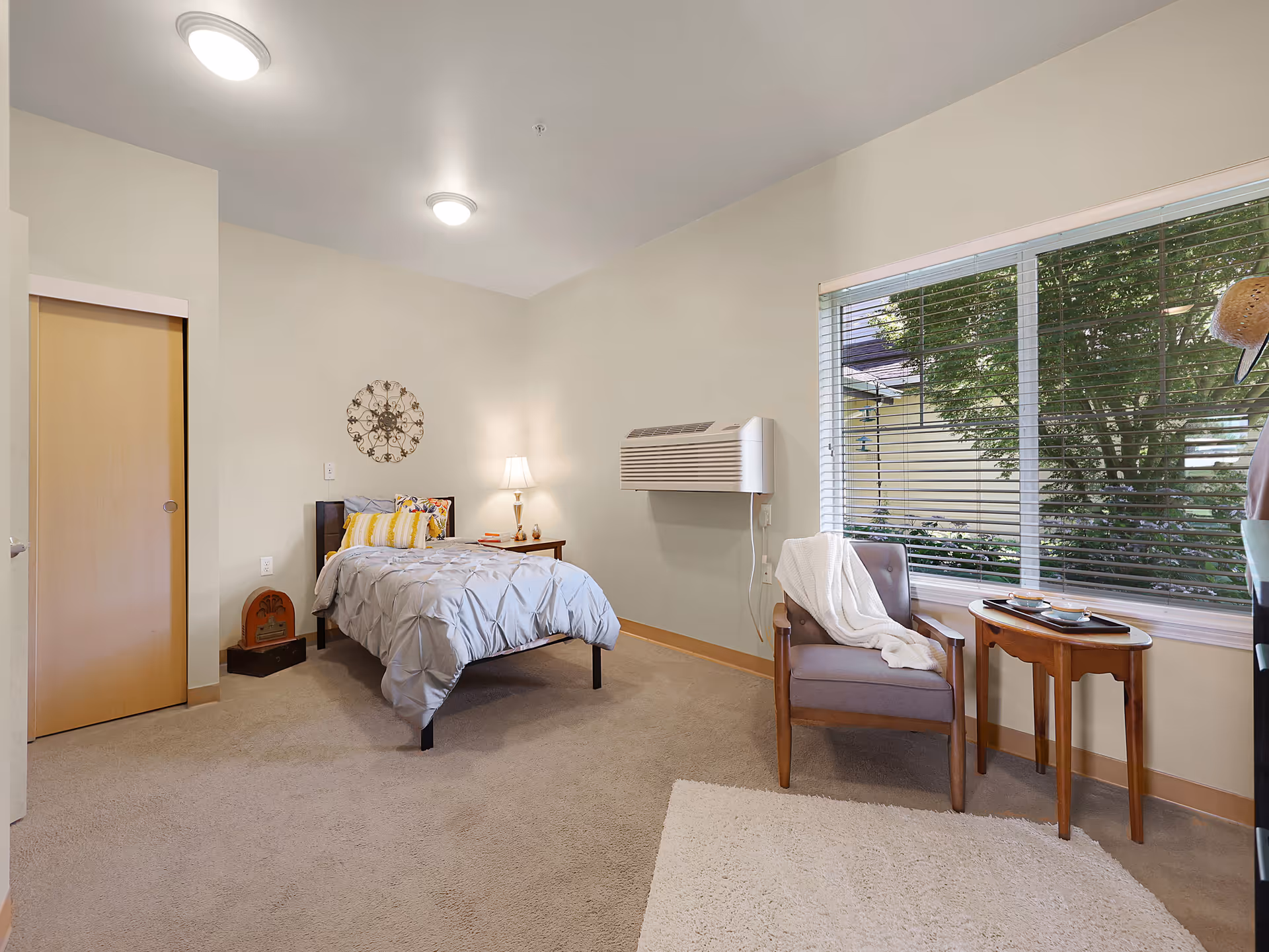 A cozy bedroom with a single bed dressed in gray bedding and yellow accent pillows. Next to the bed is a wooden nightstand with a lamp and some books. A decorative wall piece hangs above the bed. There is a wooden chair with a white blanket draped over it beside a small wooden table near a large window with blinds, showing greenery outside. The room has beige walls, carpeted floor, and a wall-mounted air conditioning unit.