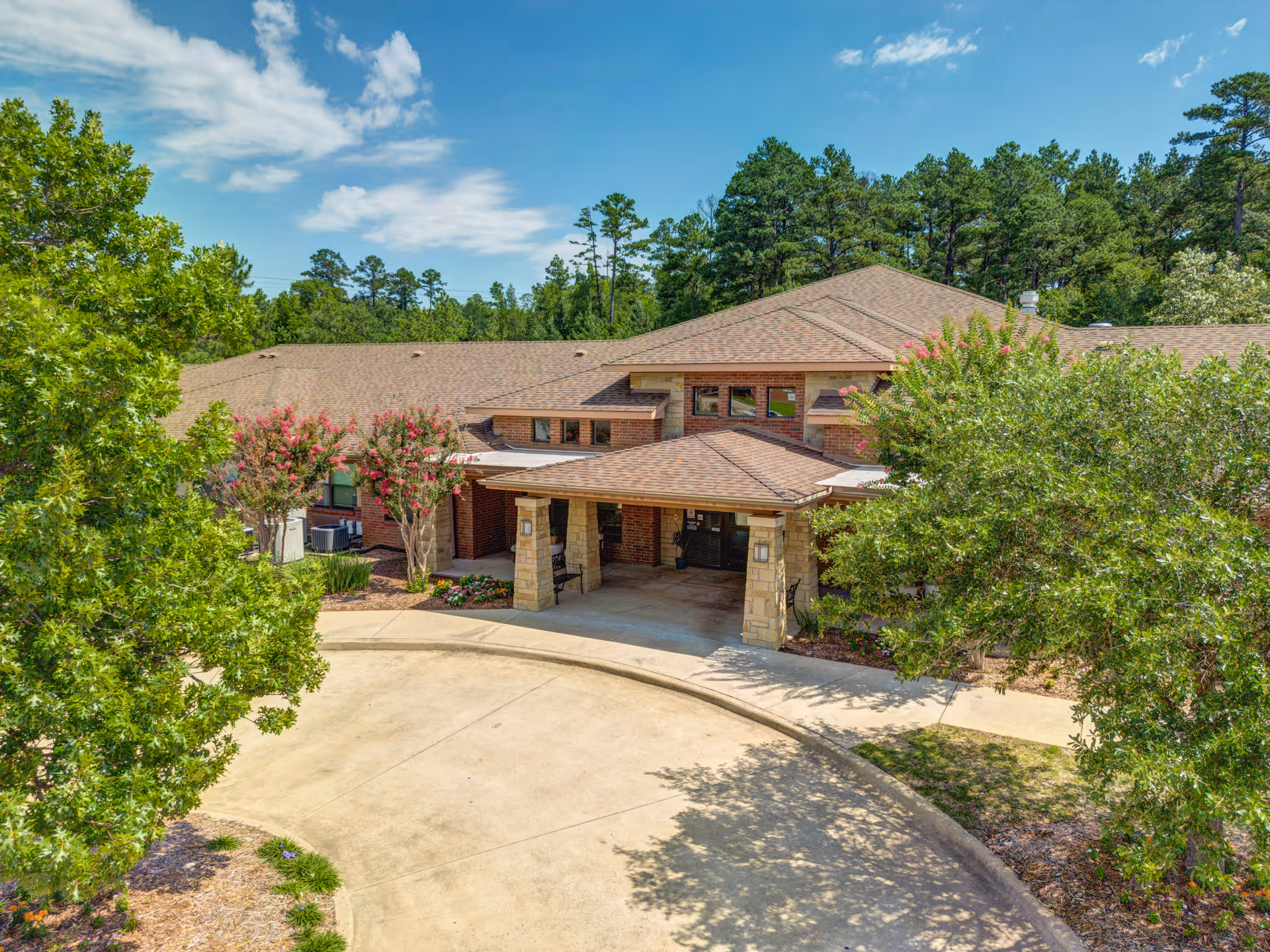 Front exterior view of Oak Hills Terrace Memory Care facility showing a single-story brick building with a covered entrance, surrounded by trees and landscaping under a blue sky with some clouds.