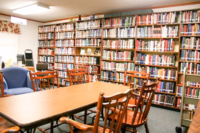 A cozy library room with wooden bookshelves filled with books lining the walls. In the center, there is a rectangular wooden table surrounded by wooden chairs with armrests. A blue upholstered armchair is visible on the left side near a window with floral curtains. The room is well-lit with ceiling lights.