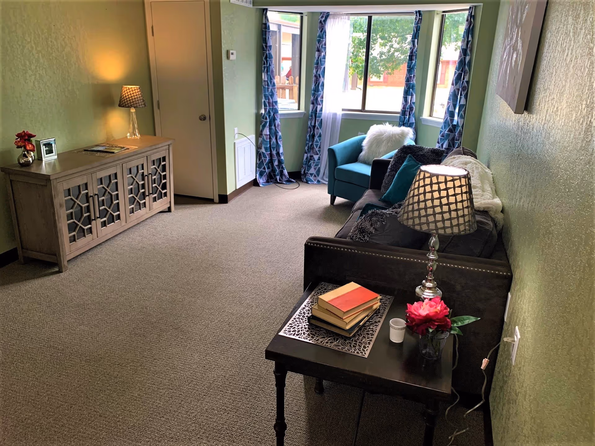 A cozy living room with green walls and carpeted floor. There is a dark sofa with pillows and a white throw blanket, a small side table with a lamp, books, and a flower vase. A teal armchair with a white fluffy pillow is near a window with blue patterned curtains. A wooden cabinet with decorative doors and a lamp is against the opposite wall.