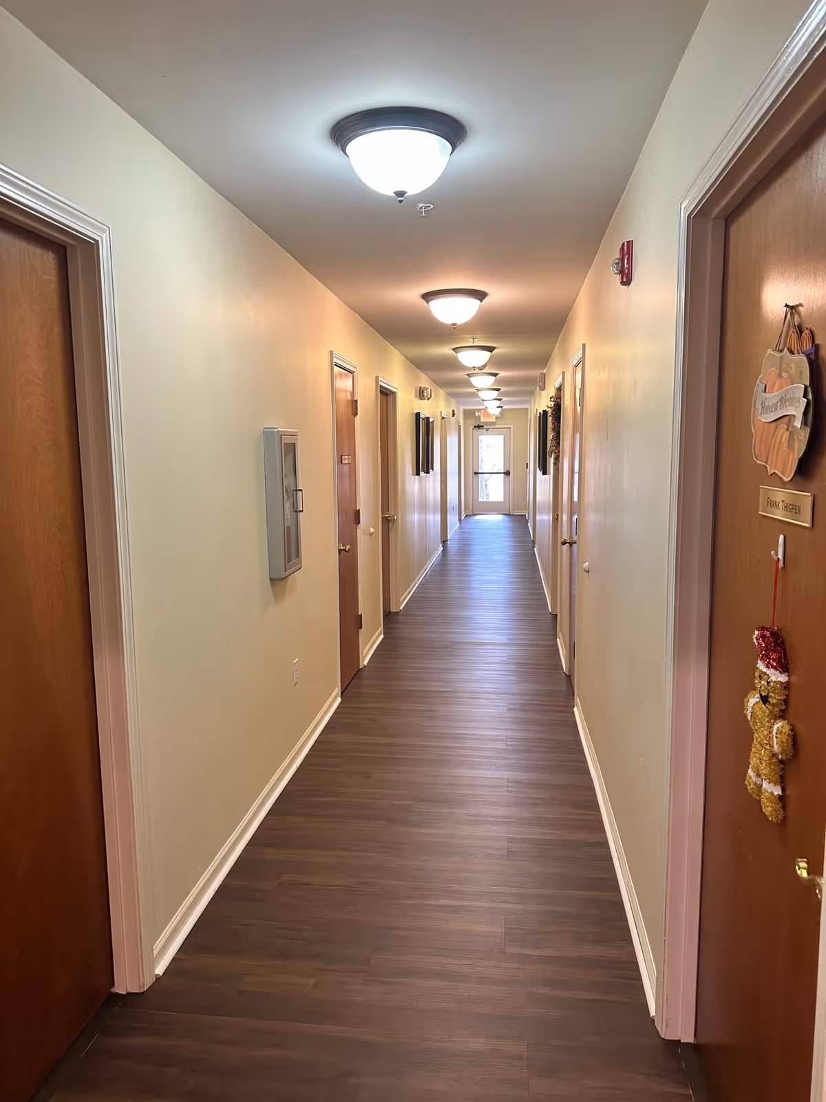 Long interior hallway in a senior living facility with wood-look flooring, multiple apartment doors and ceiling lights leading to a distant exit.