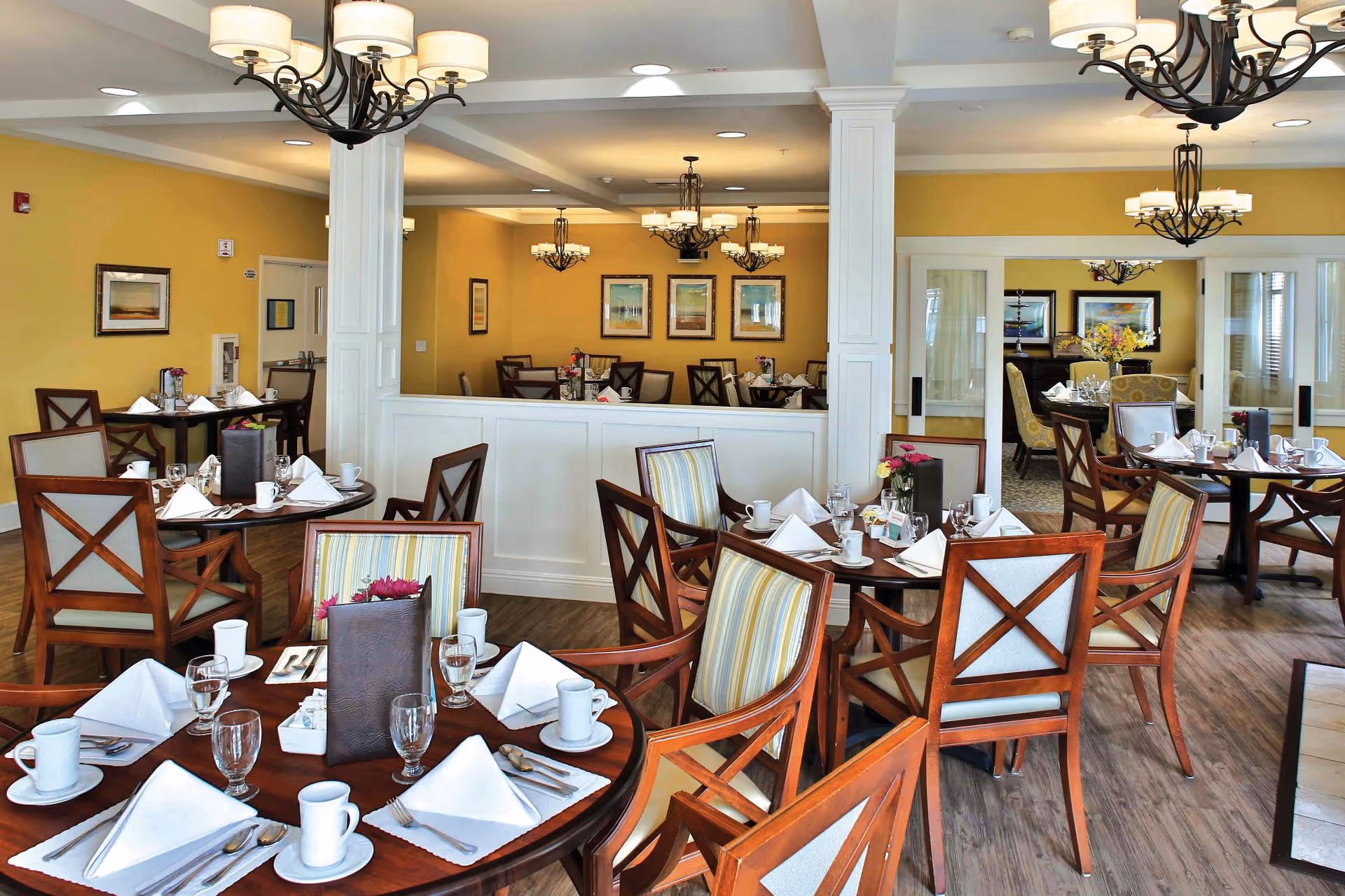 A bright and elegant dining room with multiple round wooden tables set with white napkins, cups, glasses, and silverware. The room features wooden chairs with striped cushions, yellow walls adorned with framed artwork, and several chandeliers hanging from the ceiling. There is a white partition with columns separating two sections of the dining area.