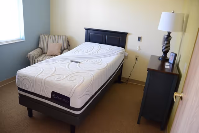 A simple bedroom with a single bed featuring a white patterned mattress, a striped armchair with a beige pillow near a window, and a dark wooden nightstand with a lamp and a framed photo on it. The walls are light-colored and the floor is carpeted.