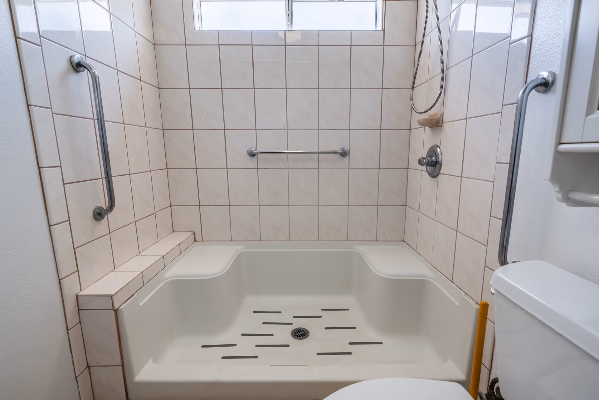 A tiled shower area with built-in seats on both sides, grab bars on the walls for support, a handheld showerhead, and a soap holder. The shower floor has anti-slip strips and a drain in the center. Part of a toilet is visible on the right side.