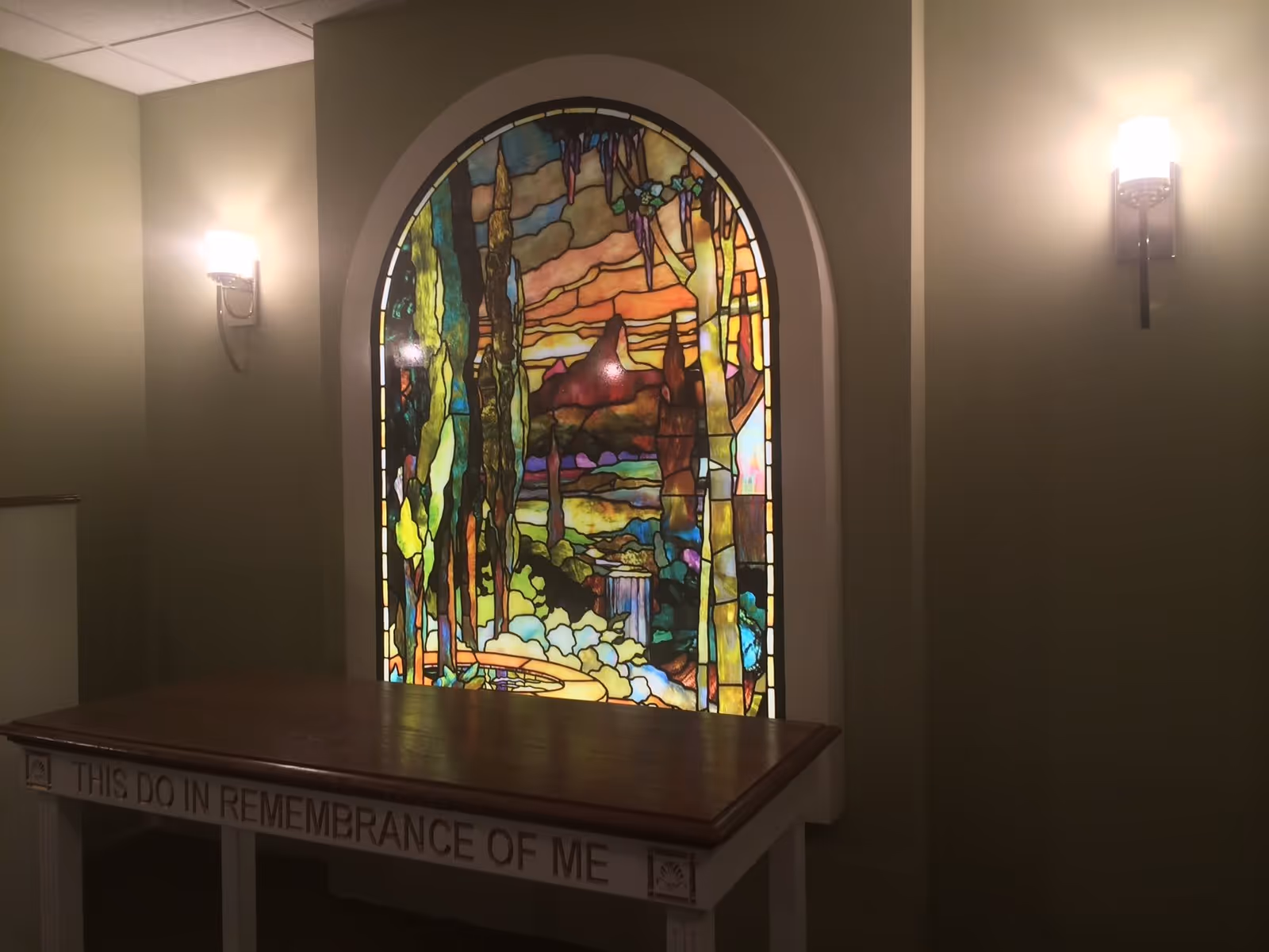 A stained glass window with a colorful nature scene featuring trees, a waterfall, and a sunset, set in an arched frame on a beige wall. Below the window is a wooden table with the inscription 'THIS DO IN REMEMBRANCE OF ME'. Two wall-mounted light fixtures with frosted glass shades illuminate the area.