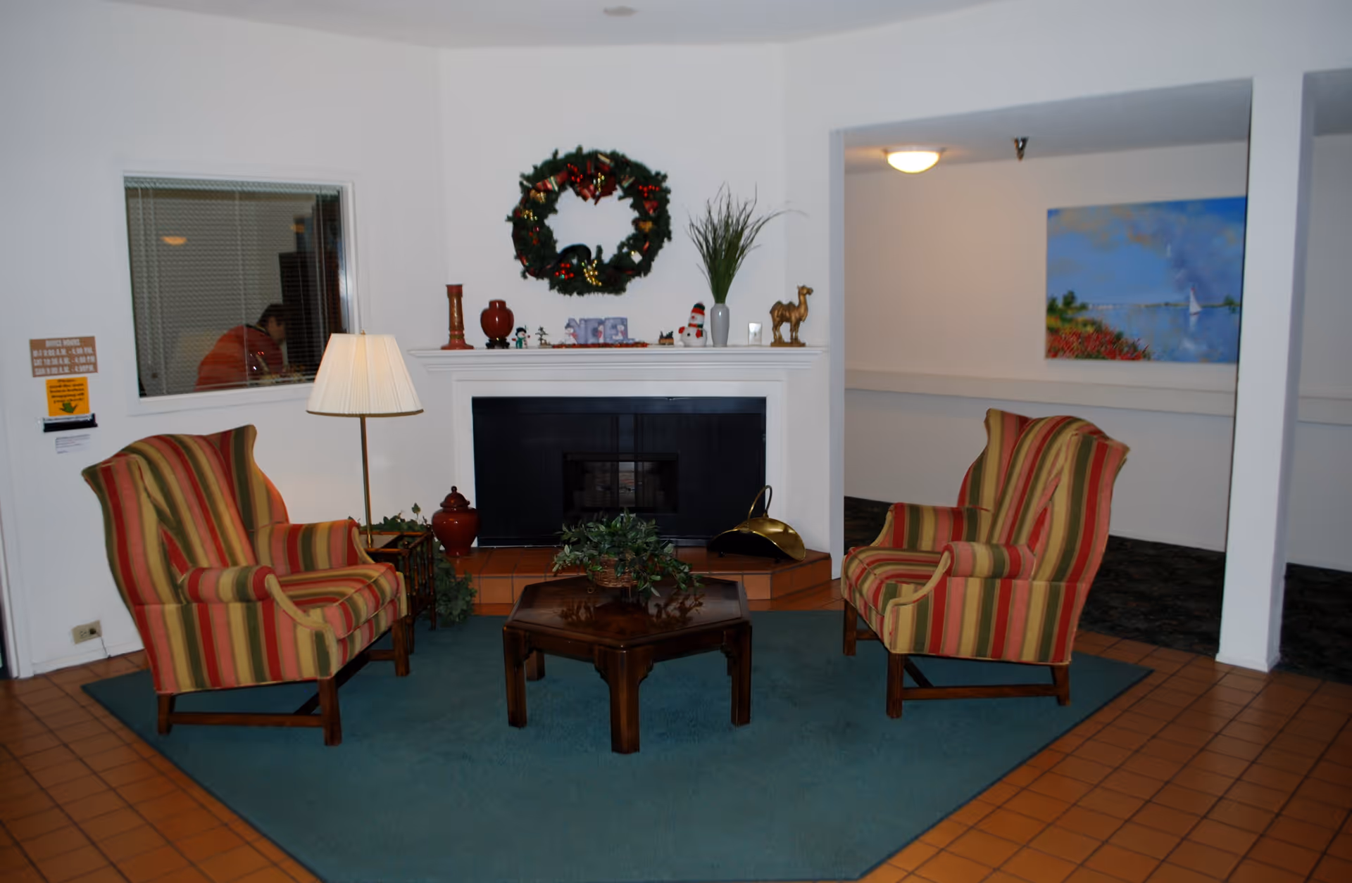 A cozy sitting area with two striped armchairs facing each other across a wooden coffee table on a teal rug. Behind the chairs is a white fireplace decorated with a Christmas wreath, small figurines, a plant, and other decorative items. A floor lamp stands next to one of the chairs, and a window with blinds is visible on the left wall. The floor is tiled in a reddish-brown color.