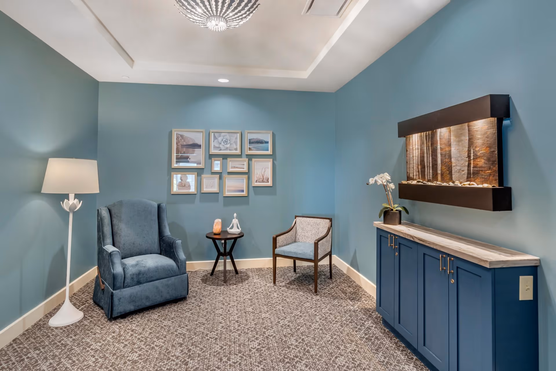A cozy sitting area with blue walls featuring a blue upholstered armchair and a wooden chair with blue cushions. Between the chairs is a small round wooden table with decorative items. On the right side, there is a blue cabinet with a marble top and a wall-mounted water feature above it. A white floor lamp stands to the left, and a collection of framed pictures is arranged on the wall behind the chairs.