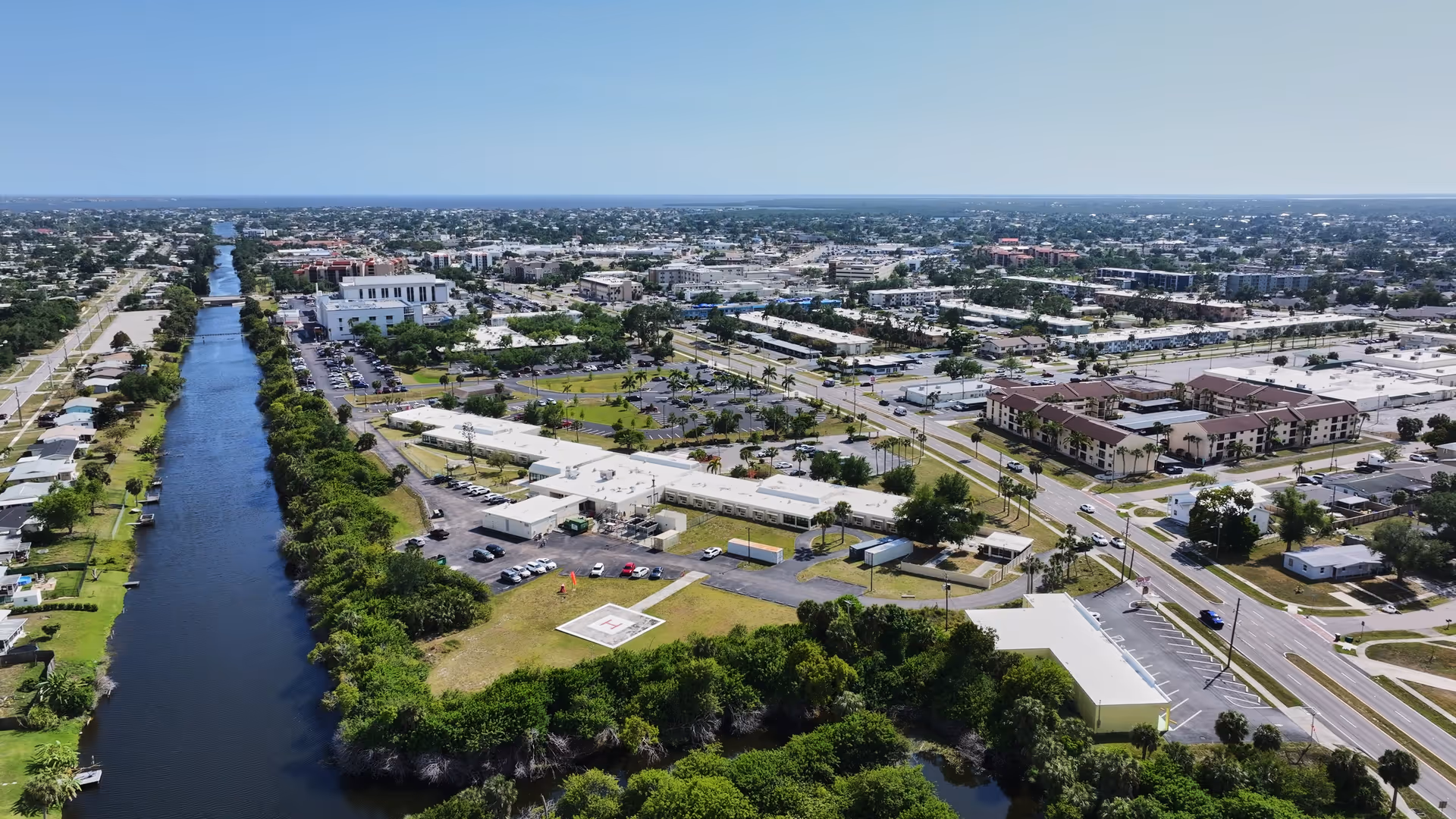 Aerial view of a suburban area featuring a long canal running parallel to a road lined with houses and trees. The image shows multiple buildings, parking lots, and roads with vehicles. There is a helipad visible near the center of the image, surrounded by greenery and several low-rise buildings. The sky is clear and blue.