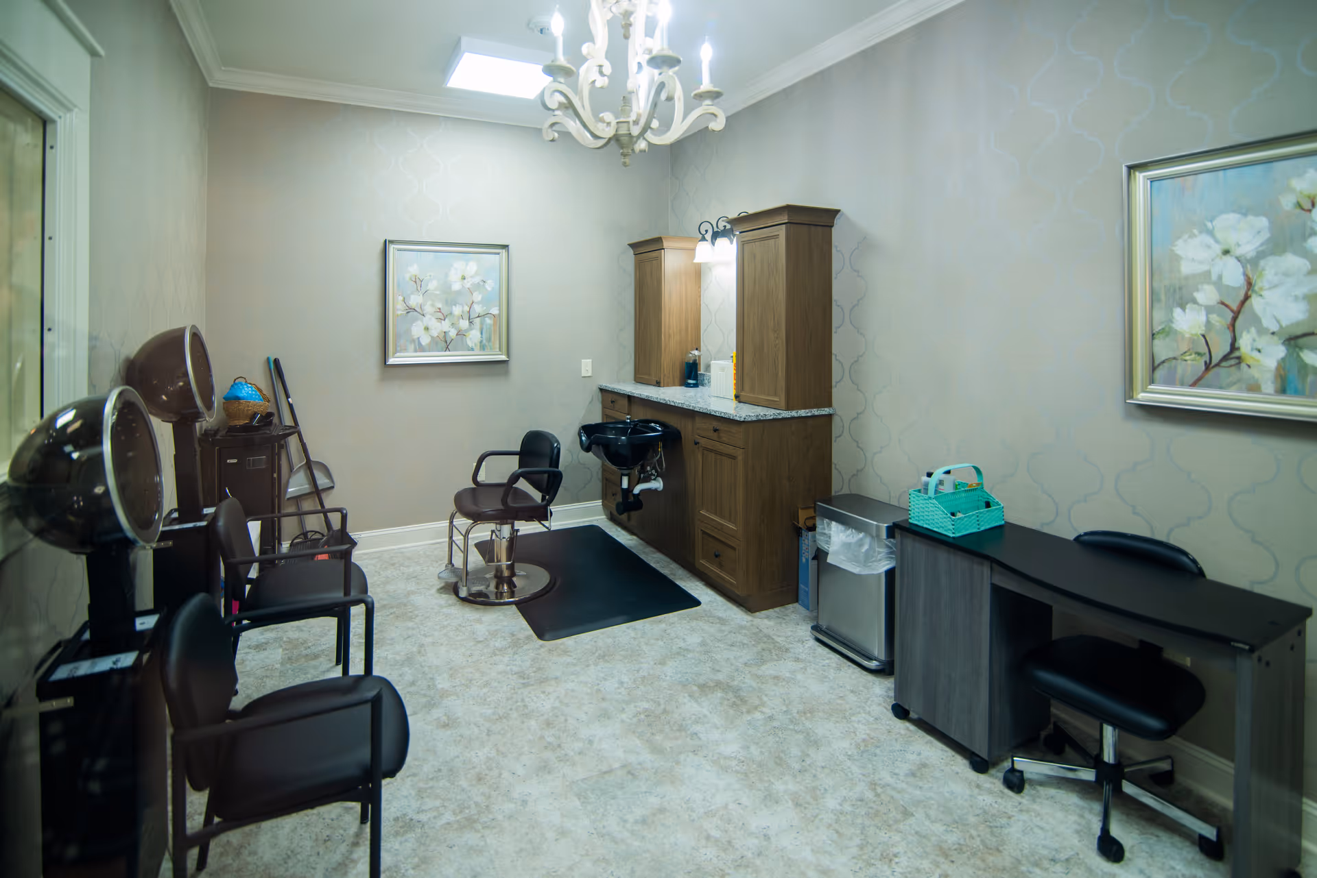 Interior view of a hair salon area in a senior living facility featuring salon chairs, hair drying stations, a hair washing sink, wooden cabinets with countertop, a small desk with a chair, and floral artwork on the walls. The room has soft lighting and a chandelier hanging from the ceiling.