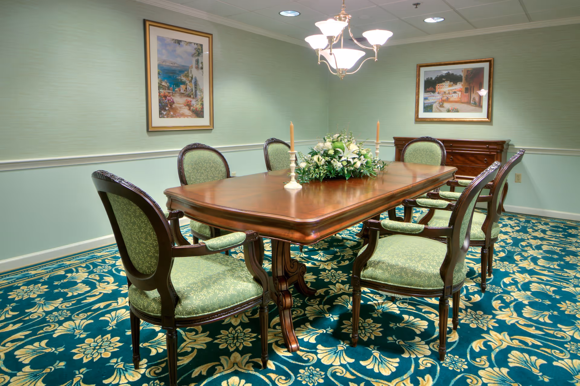 A formal dining room with a wooden dining table surrounded by six green upholstered chairs. The table is decorated with a floral centerpiece and two candlesticks. The room has green walls with white trim, two framed paintings, a chandelier overhead, and a patterned carpet in shades of green and beige.