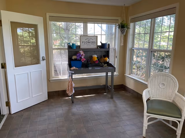 A bright room with large windows showing outdoor greenery. Inside, there is a white door on the left, a black potting bench with various colorful artificial flowers and gardening tools, and a white wicker chair with a green cushion on the right. The floor is tiled with square tiles.