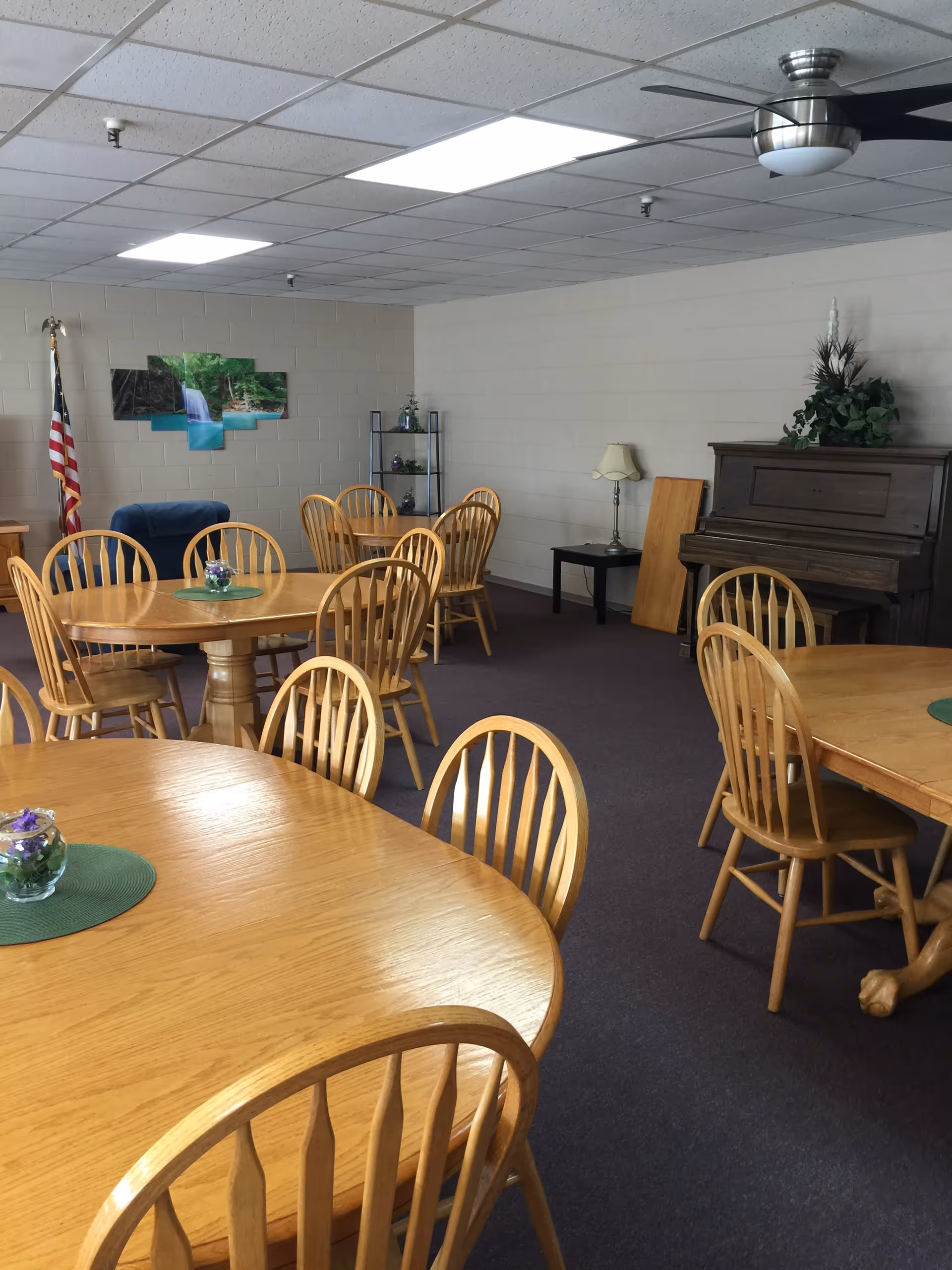 A communal room with several wooden tables and chairs arranged on a carpeted floor. The room has a white drop ceiling with fluorescent lights and a ceiling fan. There is a piano against one wall with a plant on top, a small side table with a lamp, a blue armchair, an American flag, and a wall decoration featuring a waterfall scene.