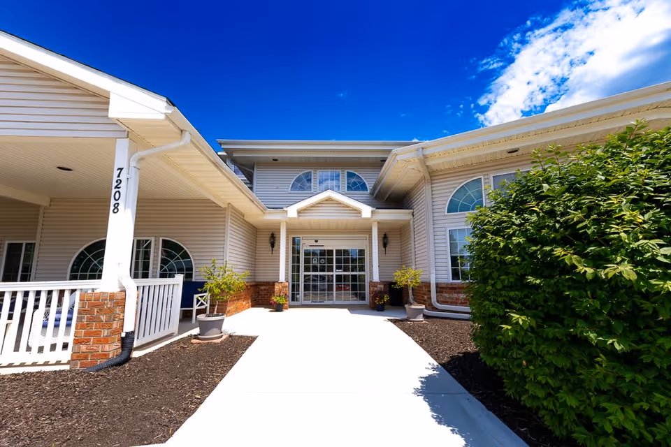 Front exterior view of Bailey Pointe Assisted Living at Van Dorn showing the entrance with a covered porch, large windows with decorative arches, potted plants, and a clear blue sky above.