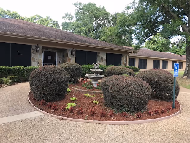 Exterior view of a single-story building with a brown roof and stone facade, surrounded by neatly trimmed bushes and a small garden with a multi-tiered fountain in the center. A paved walkway curves around the garden, and a blue handicap parking sign is visible on the right side. Large trees are in the background.