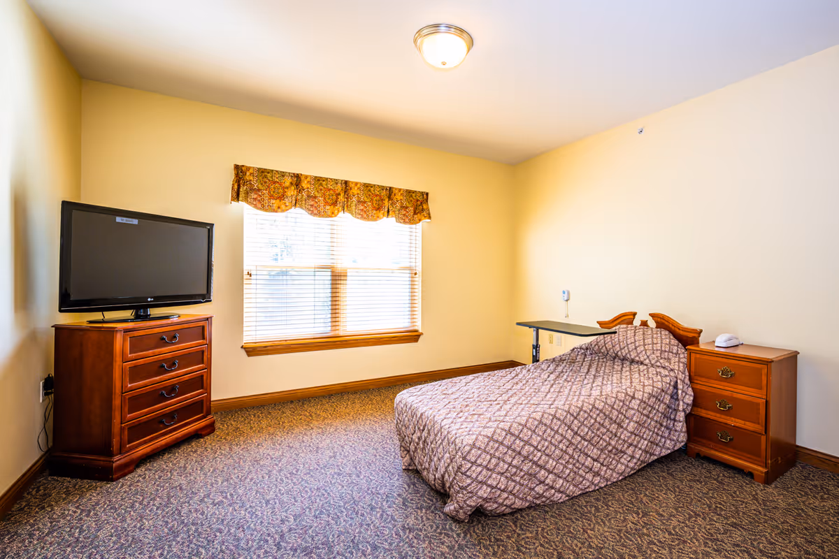 A simple bedroom with a single bed covered in a patterned bedspread, a wooden nightstand with a telephone, a wooden dresser with a flat-screen TV on top, a window with blinds and a floral valance, and beige walls with a ceiling light fixture.