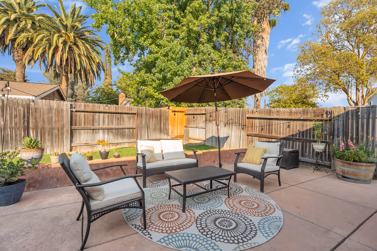 Outdoor patio area with cushioned wicker chairs and a loveseat arranged around a rectangular coffee table on a circular patterned rug. A large brown umbrella provides shade. The patio is enclosed by a wooden fence with various potted plants and trees visible in the background under a clear blue sky.