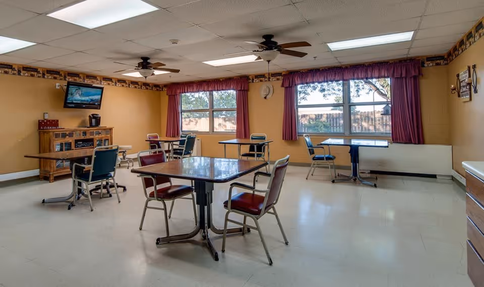 A dining room with several tables and chairs arranged around the room. The walls are painted yellow with a decorative border near the ceiling. Two large windows with red curtains let in natural light. A wooden cabinet with a coffee maker and a wall-mounted TV are visible. The ceiling has fluorescent lights and two ceiling fans.