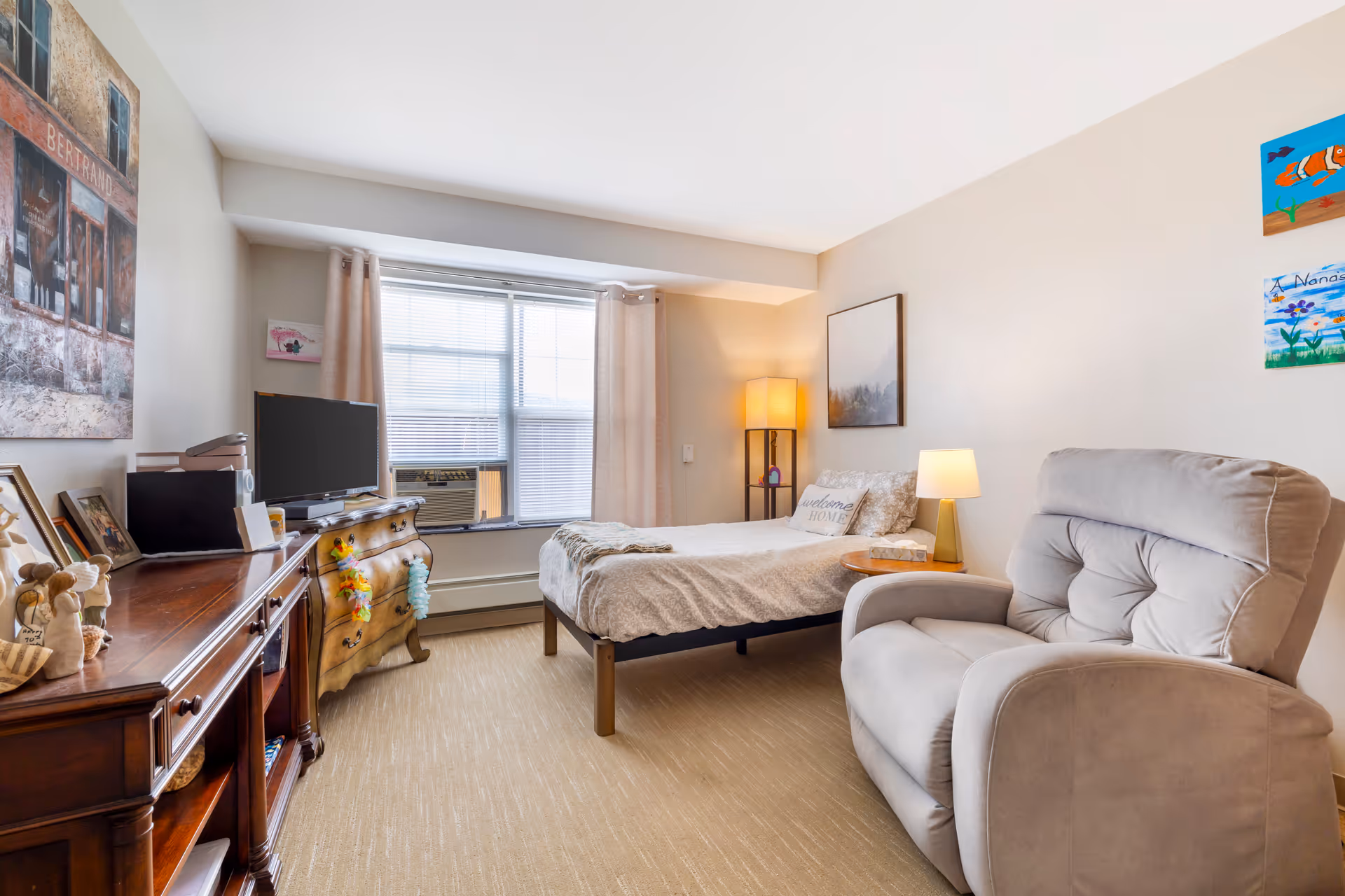 Sunny studio-style bedroom with a bed, recliner, dresser and a TV by a large window.