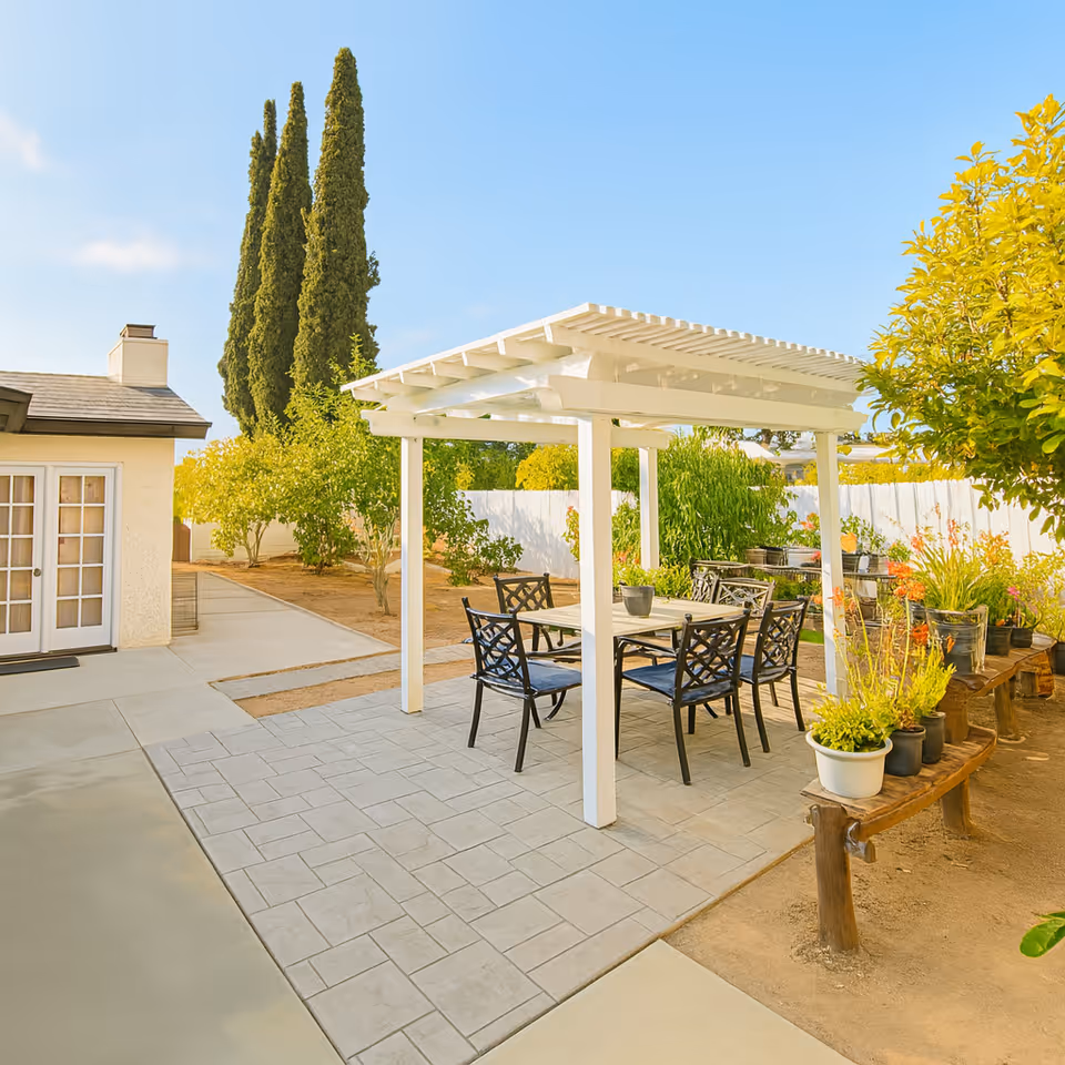 Outdoor patio area with a white pergola covering a table and six chairs. Surrounding the patio are various potted plants and trees, with a white fence and a building with French doors in the background under a clear blue sky.