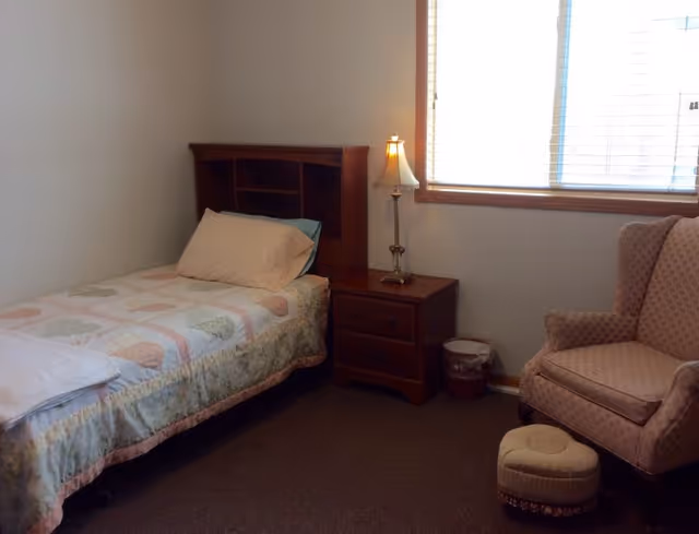 A small bedroom with a single bed covered with a pastel-colored quilt and a pillow. Next to the bed is a wooden nightstand with two drawers and a decorative lamp on top. There is a window with blinds letting in natural light. To the right of the nightstand is a cushioned armchair with a matching footstool. The room has plain white walls and a carpeted floor.
