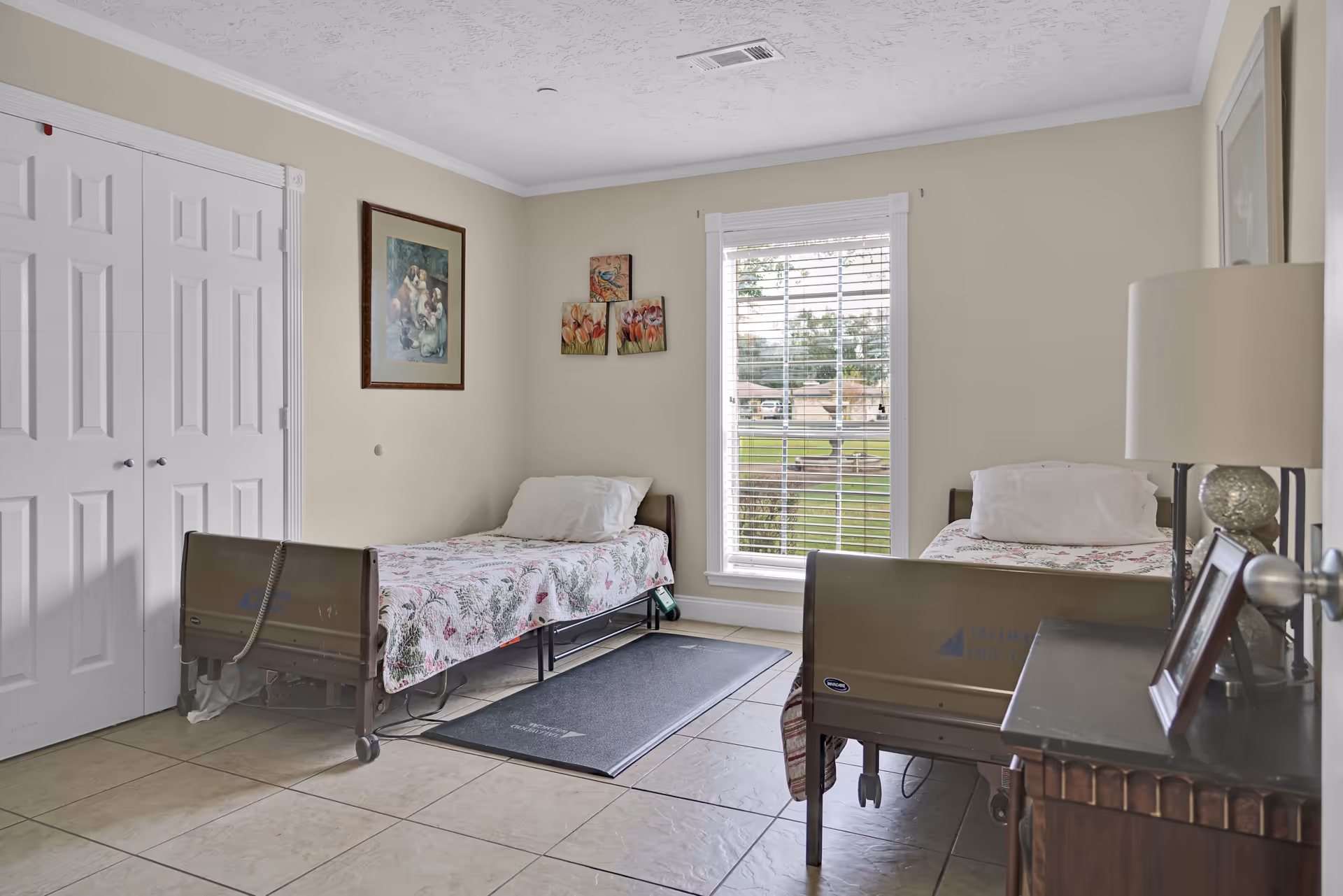 A simple, clean bedroom with two hospital-style beds, each with floral bedspreads and white pillows. The room has beige walls with framed artwork, a large window with white blinds letting in natural light, a dark wooden nightstand with a lamp and picture frames, and a tiled floor with a black mat between the beds.