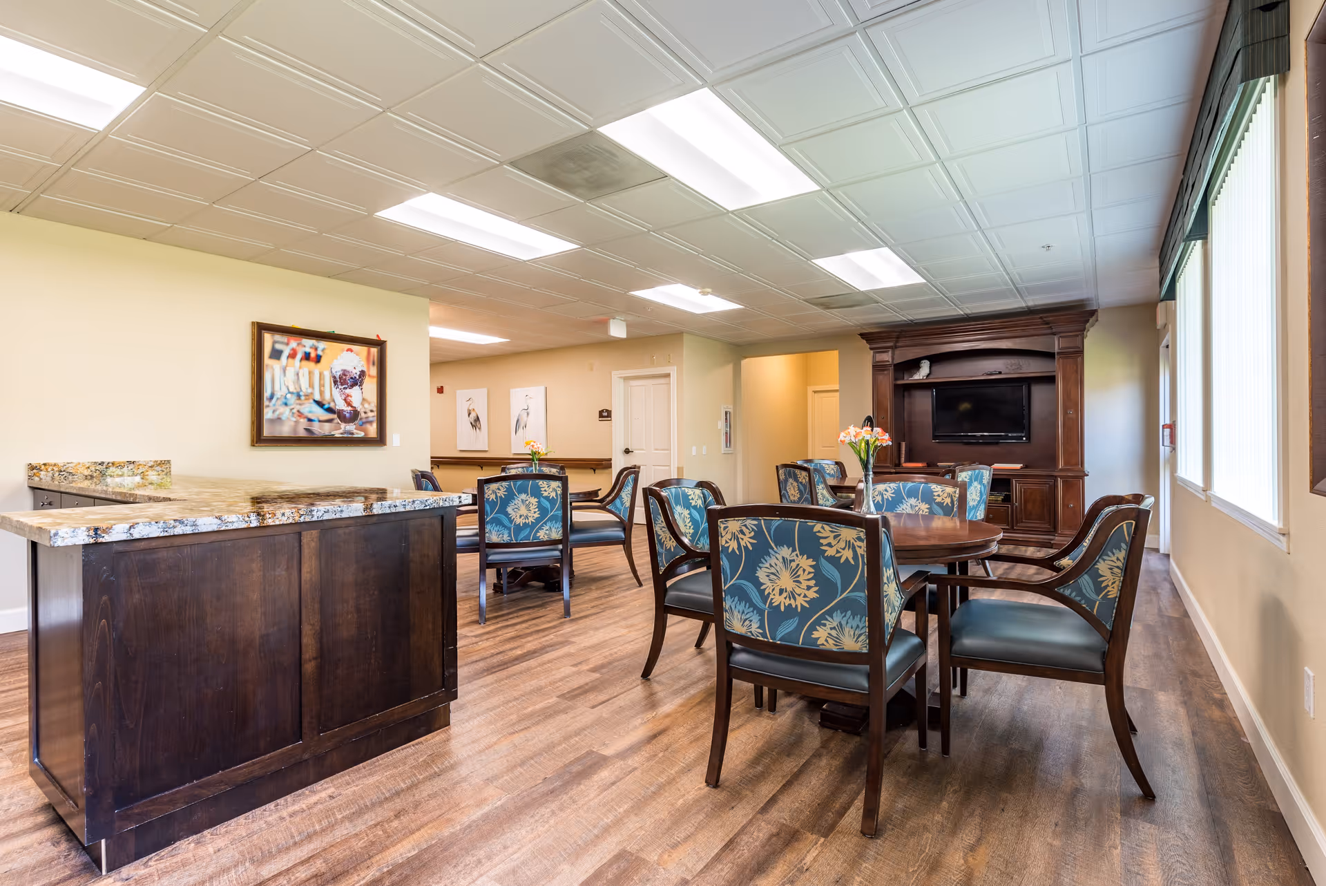 A well-lit common area with wooden flooring, featuring a dark wood island with a granite countertop on the left, and several round tables surrounded by chairs with blue floral upholstery. A large wooden entertainment center with a TV is against the far wall, and windows with blinds line the right side of the room. Artwork is displayed on the walls.