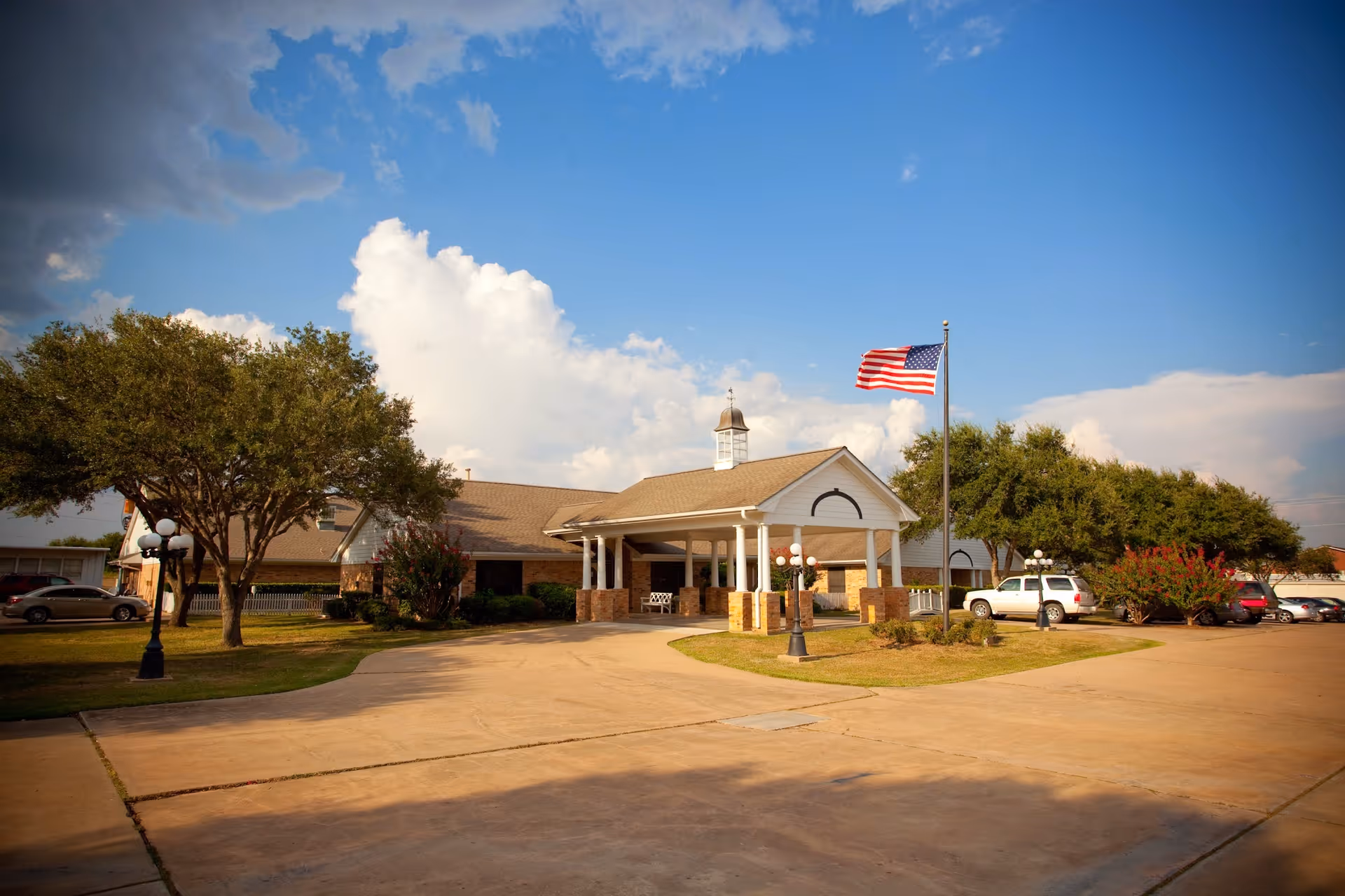 Front entrance of a single-story senior living facility with a porte-cochere, American flag, driveway, parked cars and trees under a blue sky.