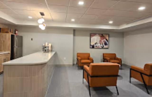 A modern interior room with a long kitchen island on the left side featuring a refrigerator and coffee machine. On the right side, there are four orange armchairs arranged around a small table. The walls are light gray, and a framed picture hangs on the far wall. The ceiling has recessed lighting and three hanging pendant lights above the island.