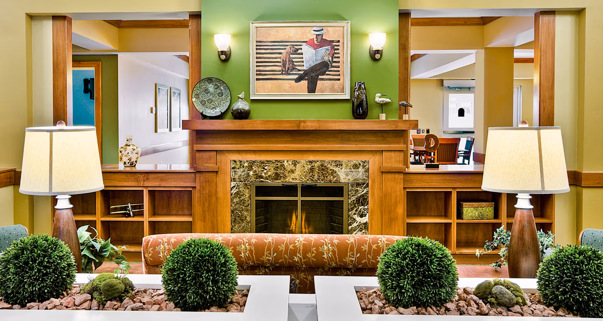 A cozy living room area featuring a wooden fireplace with a marble surround, decorated with vases and a painting of a person reading on a bench with a dog. Two table lamps with beige shades flank the fireplace, and there are green plants and bushes in the foreground. The room has warm yellow and green walls with wooden trim and built-in shelves.