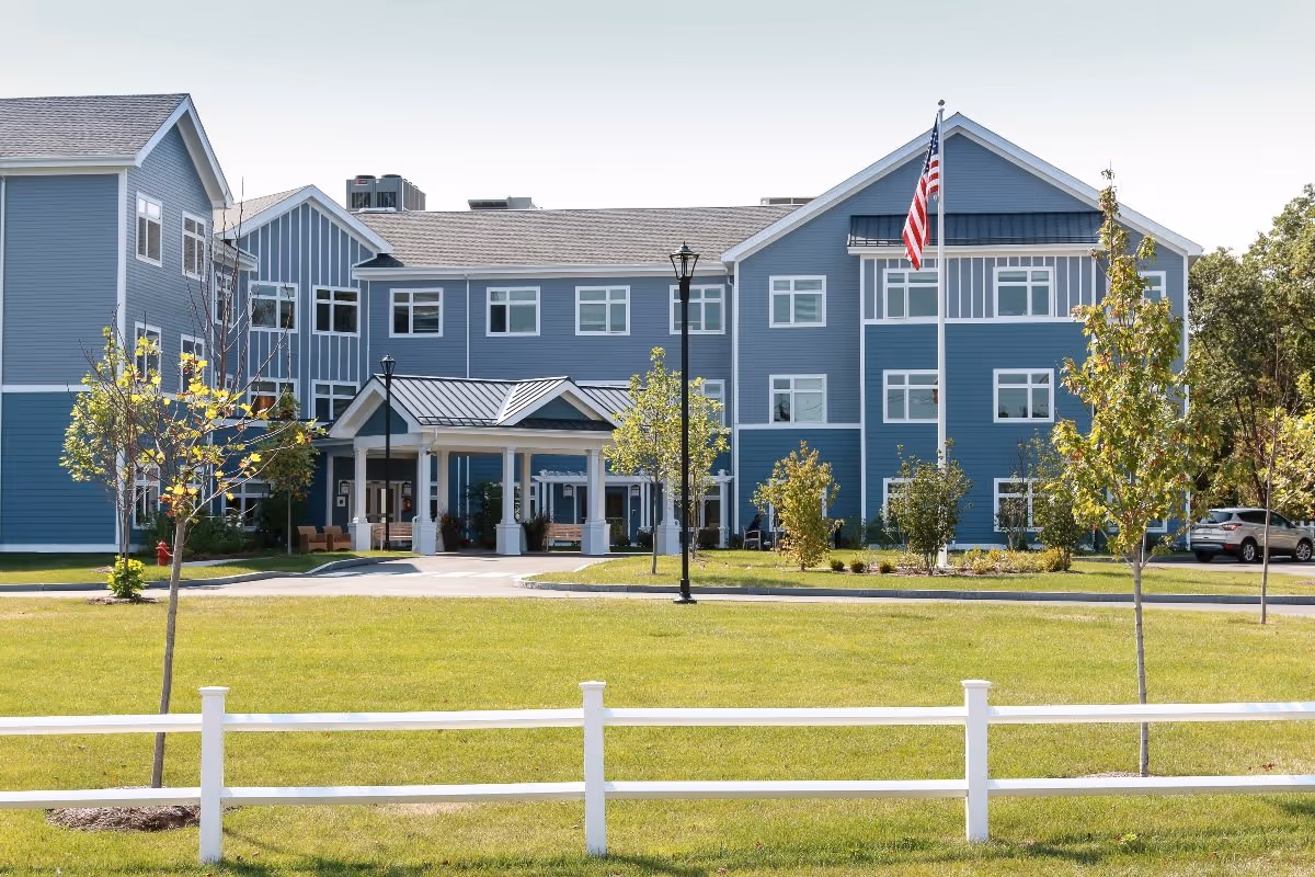Front exterior view of a large three-story blue senior living facility building with white trim, a covered entrance, an American flag on a flagpole, young trees, and a white fence in the foreground under a clear sky.