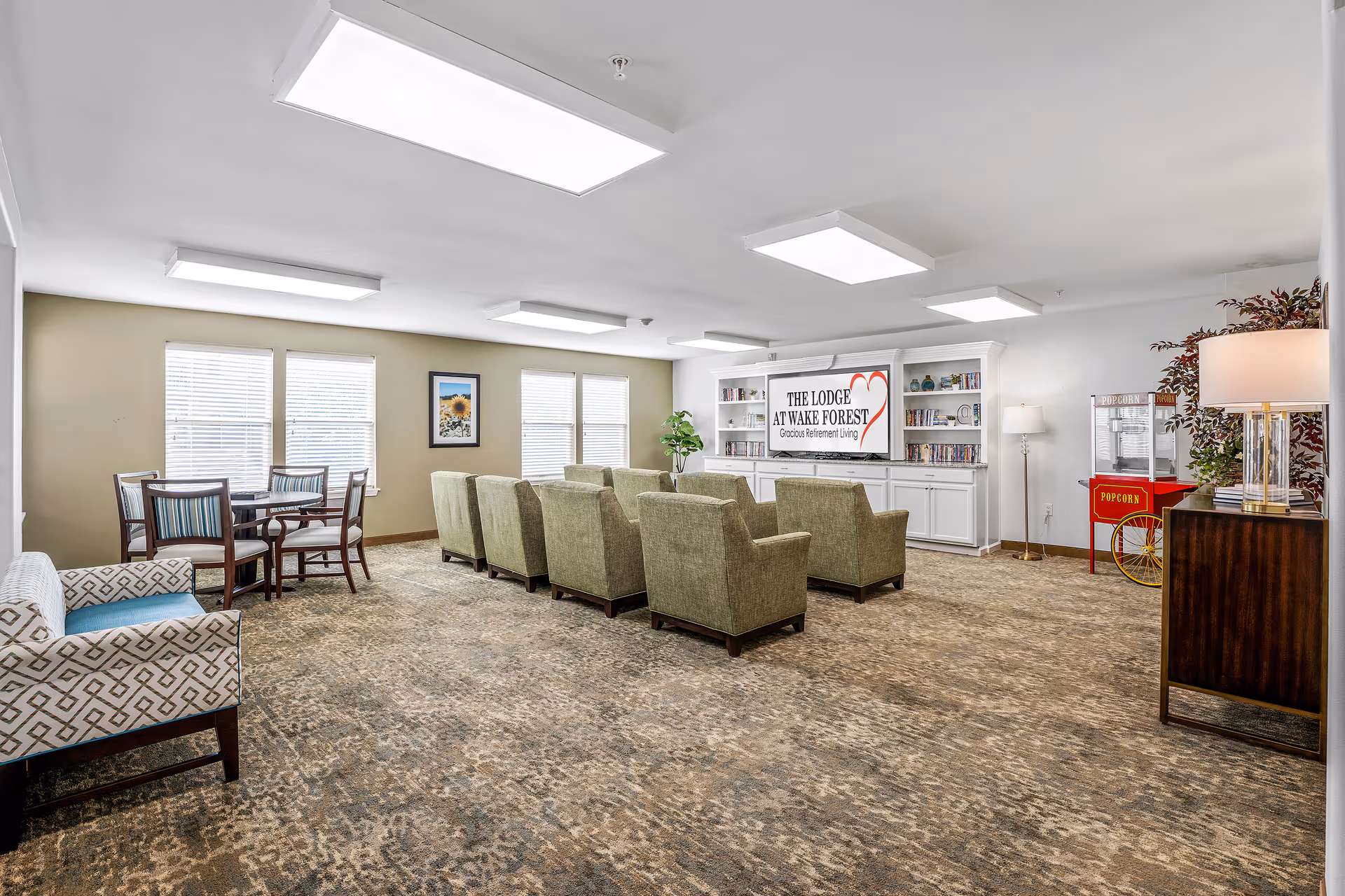 A spacious and well-lit common area in The Lodge at Wake Forest Gracious Retirement Living featuring a row of green armchairs facing a large screen with the facility's name and logo. The room includes a small round table with chairs near windows, a popcorn machine, bookshelves, and a lamp on a wooden side table.