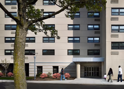 A beige multi-story senior living building with a glass entrance and people walking on the sidewalk in front.