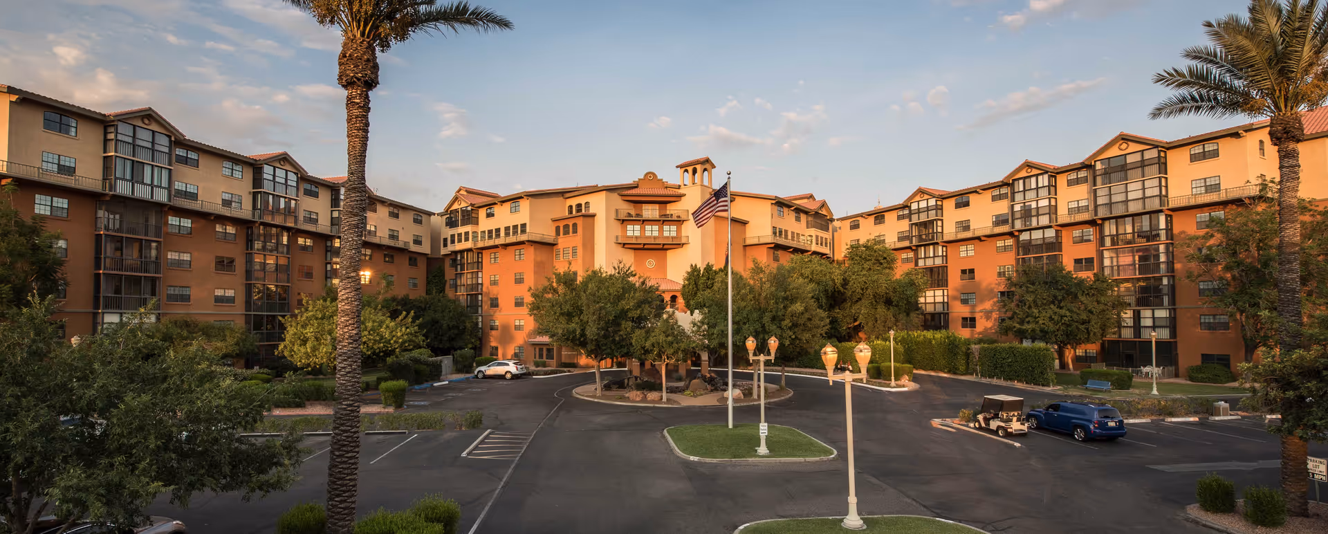 Exterior view of Freedom Plaza Care Center, a large multi-story building with a Mediterranean architectural style featuring terracotta roofs and balconies. The building is surrounded by trees and a parking lot with a few vehicles, including a golf cart. An American flag is prominently displayed on a flagpole in the center of the driveway.