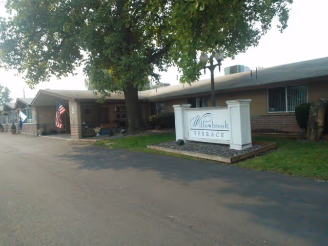 Single-story brick building entrance with a large tree, an American flag, and a sign that reads "Willowbrook Terrace".