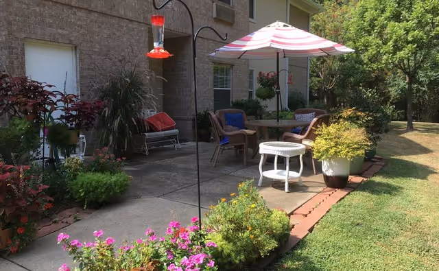 Outdoor patio area with a round table and four wicker chairs, one with a blue cushion, under a pink and white striped umbrella. The patio is surrounded by various potted plants and flowers, with a hummingbird feeder hanging from a metal hook. The building exterior is visible with a brick wall, windows, and a white door. Green grass and trees are in the background.