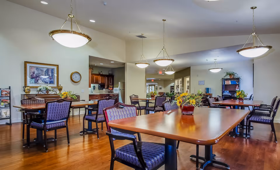 A spacious dining room in Devonshire Retirement Village with multiple wooden tables and purple cushioned chairs arranged neatly. The room features hanging pendant lights, a wooden floor, a wall clock, framed artwork, and a small kitchen area in the background. Flower arrangements are placed on the tables, and there is a bookshelf and magazine rack along the walls.